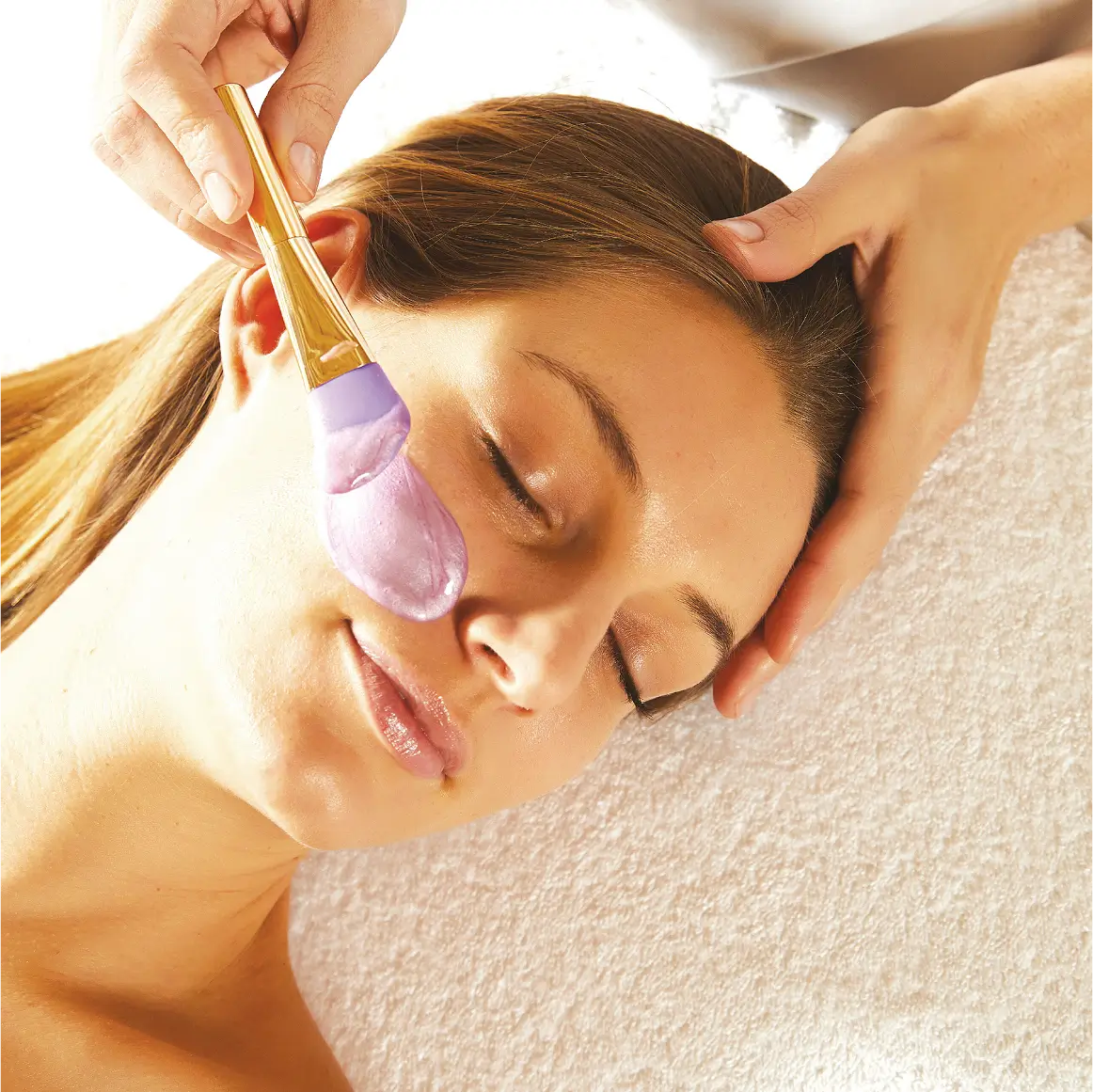 Esteticista aplicando mascarilla facial morada con pincel suave sobre el rostro de una clienta.